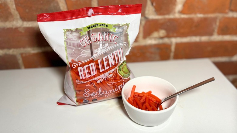 Package and white bowl of Organic Red Lentil Sedanini on a white table against a brick background