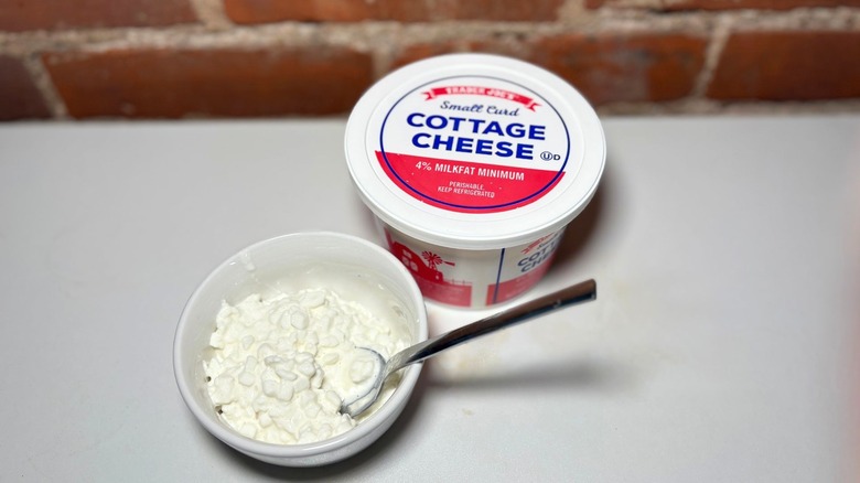 Container and white bowl of Small Curd Cottage Cheese on a white table against a brick background