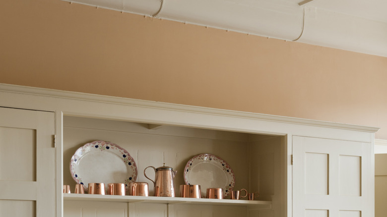 Apricot beige painted walls above a cupboard with exposed shelves with copper cups and ceramic trays.