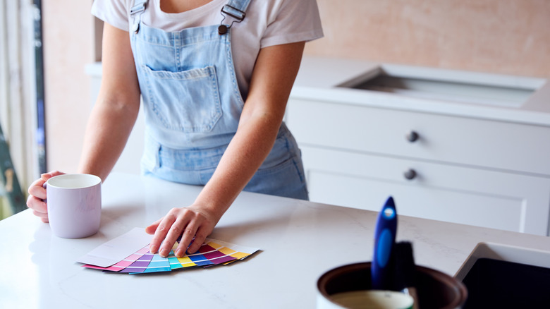 Woman in light blue overalls and a white T shirt looking at paint chips in a kitchen with a mug in one hand.