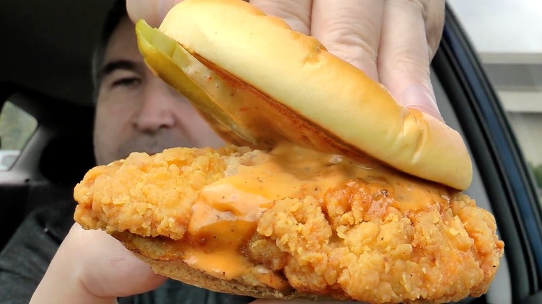 A man holds up a Jack in the Box Cluck Sandwich in a car