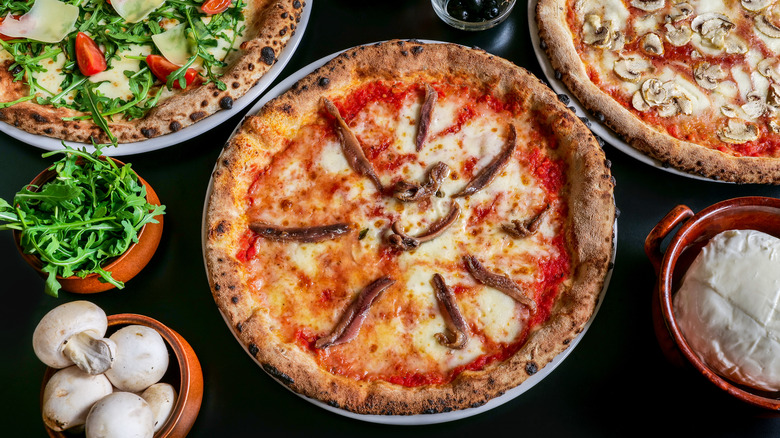 a table setting with three pizzas and garnishes of parsley, mushrooms, and peppercorns. most prominently displayed is a pizza with anchovy fillets on top of the pie
