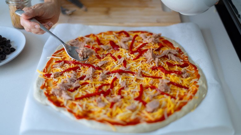 a person spoons canned tuna over an uncooked pizza with shredded cheese and red peppers