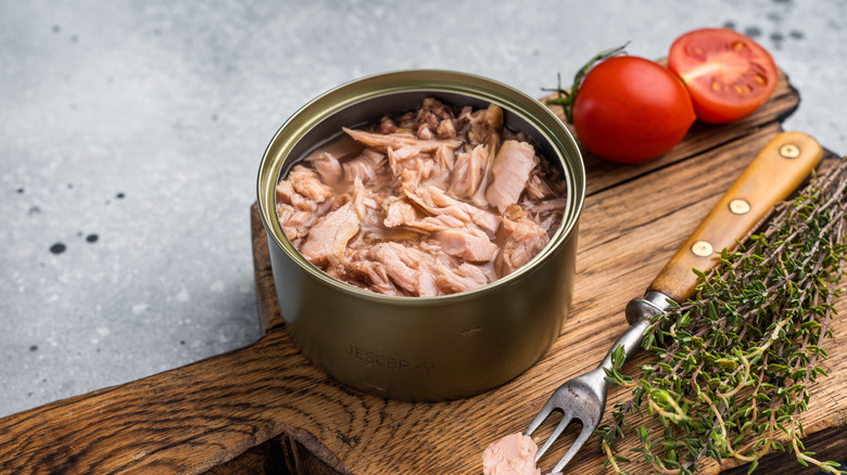 An open can of tuna with a fork, herbs, and tomatoes on a wooden cutting board