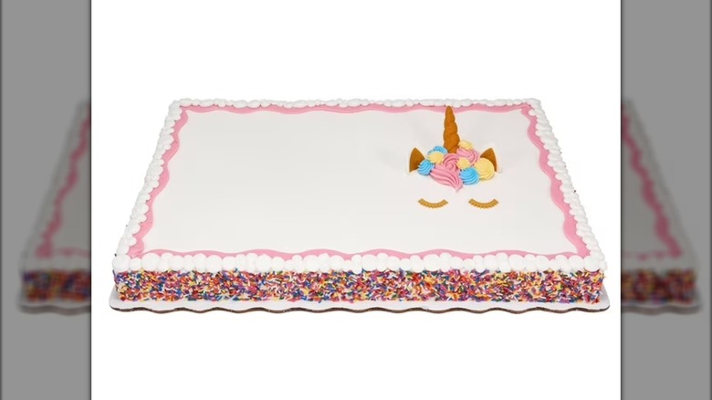 A large white sheet cake with rainbow-sprinkled sides and a unicorn face