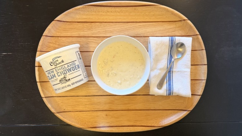 Blount's Clam Chowder in a bowl on a wooden tray next to its container, a napkin, and a spoon