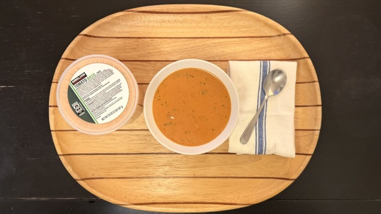 Kirkland Tomato Basil soup in a bowl on a wooden tray next to its container, a napkin, and a spoon