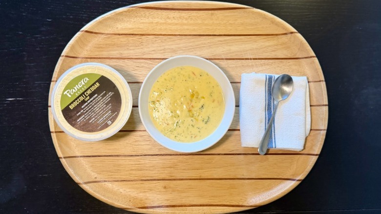 Panera Broccoli Cheddar Soup in a bowl on a wooden tray next to its container, a napkin, and a spoon
