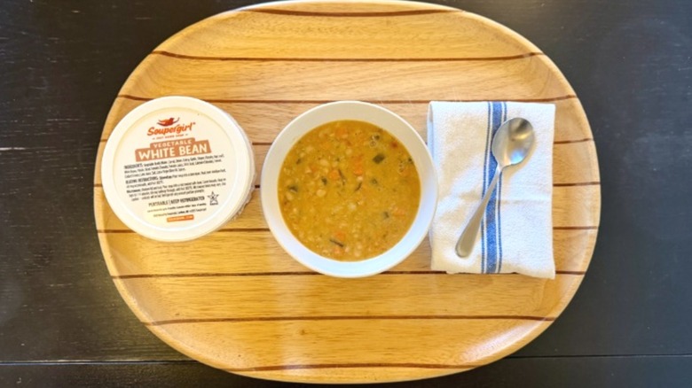 Soupergirl White Bean Soup in a bowl on a wooden tray next to its container, a napkin, and a spoon