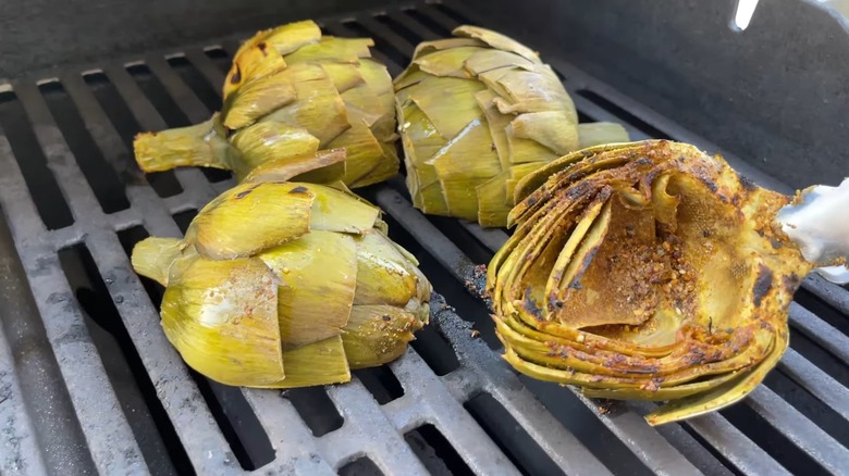 4 artichoke halves being grilled face down, with one charred face held up by tongs