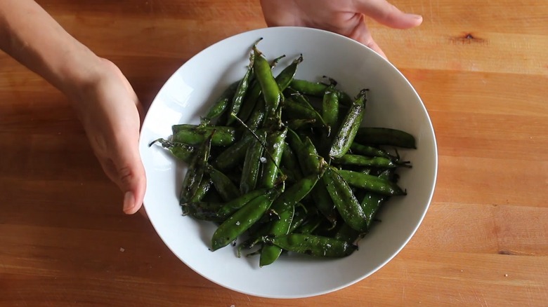 Two hands holding a white bowl of grilled snap peas, still in their pods