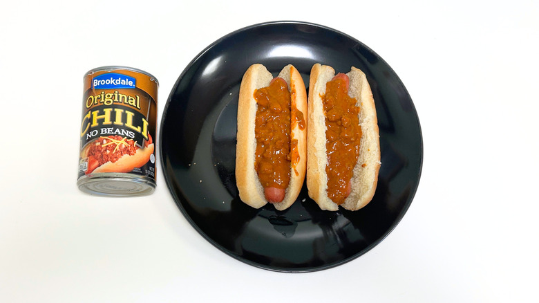 A can of Brookdale Original Chili No Beans next to a black plate with two chili hot dogs