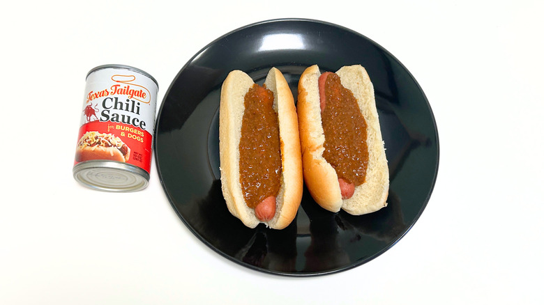A can of Texas Tailgate Chili Sauce For Burgers and Dogs next to a black plate with two chili hot dogs