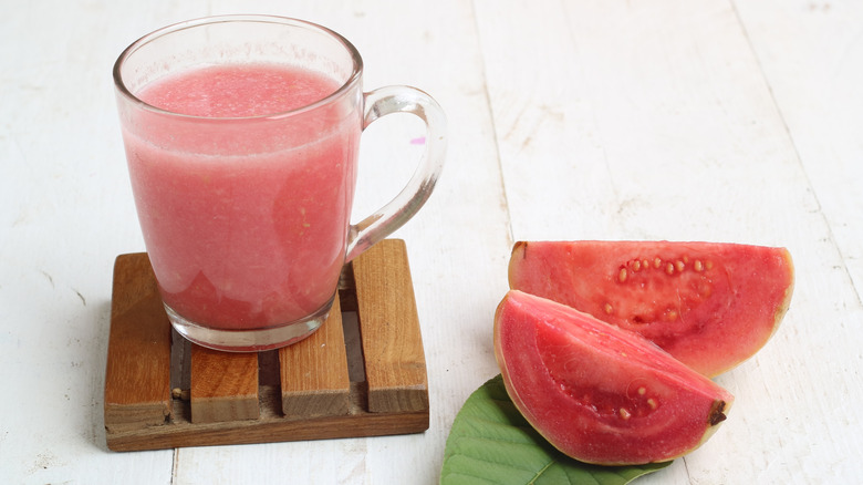A glass mug of pink guava juice on a wood coaster next to two slices of guava, all on a white table.