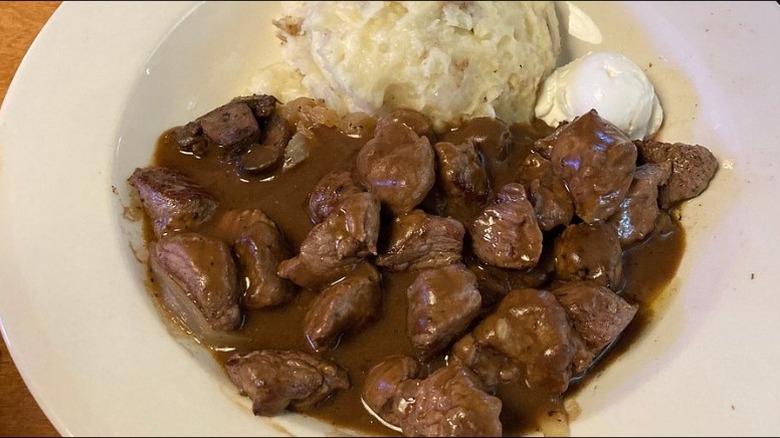 A plate with beef tips covered in gravy with a serving of mashed potatoes and a dollop of sour cream