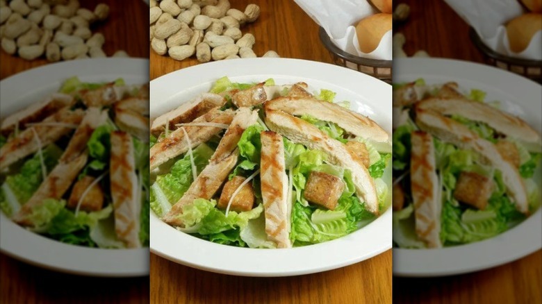 A Caesar salad with grilled chicken strips on a table, with peanuts and dinner rolls visible behind the plate