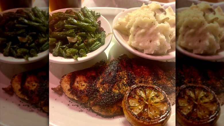 A plate with a charred chicken breast, charred lemon, and bowls of green beans and mashed potatoes