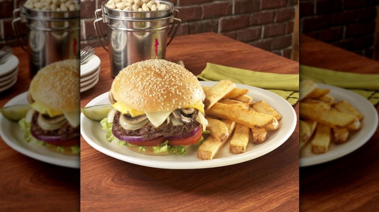 A burger topped with two slices of cheese, mushrooms, and onions on a plate with steak-cut fries, with a bucket of peanuts behind the plate