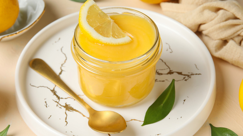A jar full of lemon curd topped with a lemon wedge on a white plate with a spoon