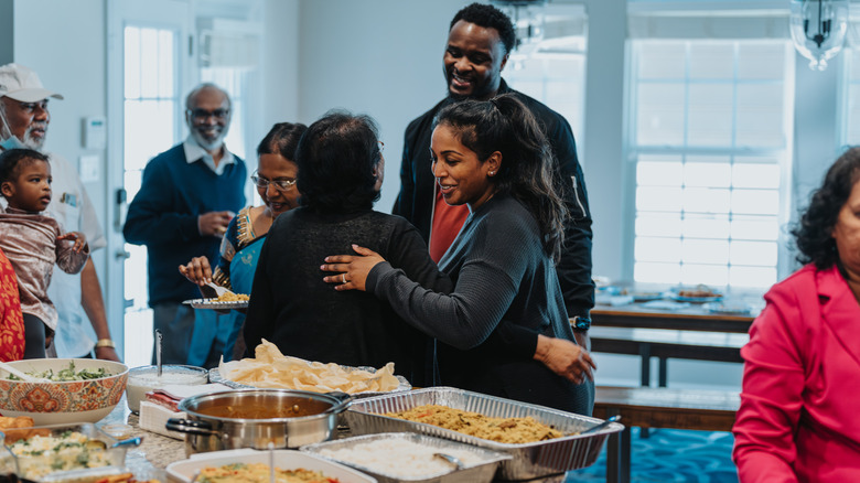 Toung and old people greet each other near a kitchen counter with bowls and trays full of food