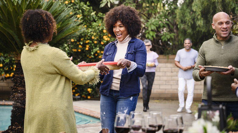 Outdoor party scene with two women greeting each other joyfully, one handing off a Tupperware container of food