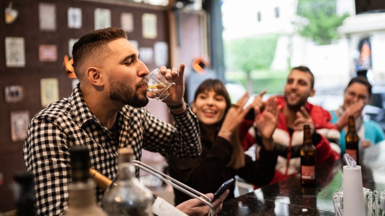 A group of four friends at a bar during daylight hours with one person drinking a glass of brown liquor