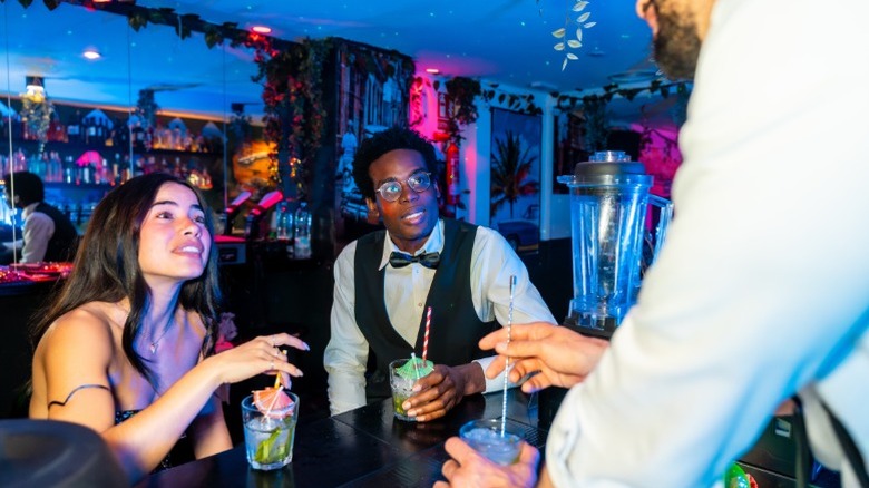 A young couple sitting at a bar with drinks and talking to the bartender at a tropical themed bar