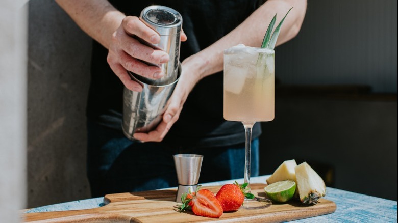 A close up of a bartender making a mocktail in a cylindrical stemmed glass sat on a wooden cutting board next to a metal jigger, strawberries, like, and pineapple