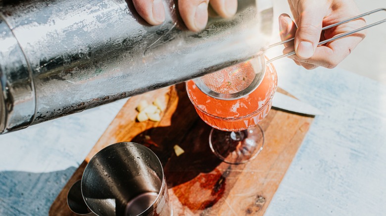 A close up of a bartender straining a cocktail into a coupe glass on a wooden cutting board next to a metal shaker tin