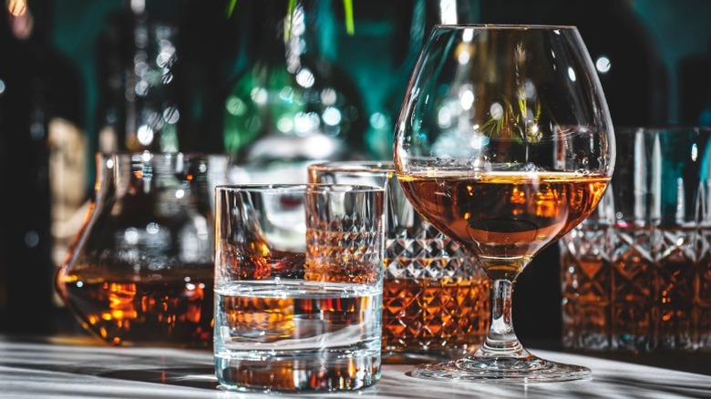 A glass of vodka and a snifter of brandy in front of crystal glassware filled with brown spirits