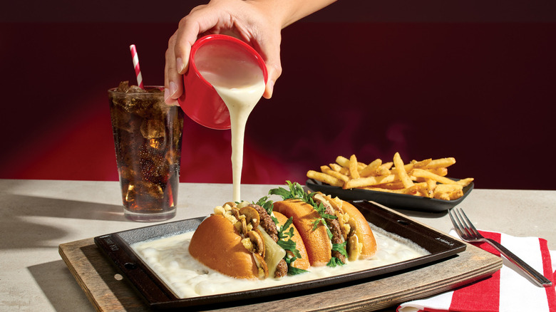 Cheese being poured from a red cup onto a meat sandwich sub cut in half, with fries and a glass of coke in the background