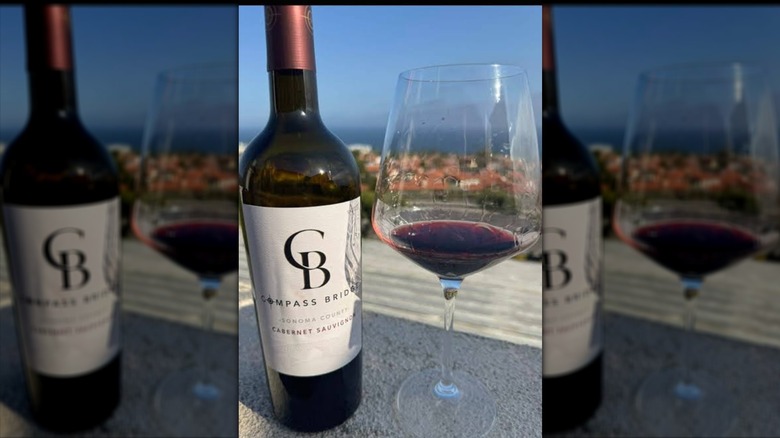 Compass Bridge wine bottle and glass with red wine in it and a view of a town and ocean in the background.
