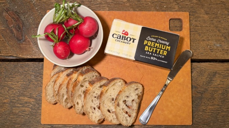 Cabot Extra Creamy Premium Butter in its packaging on a cutting board next to a butter knife, sliced baguette, and bowl of radishes