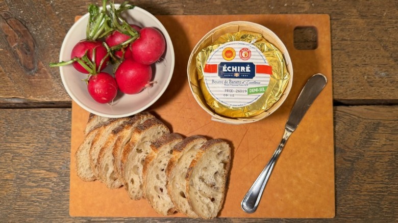 Échiré Butter in its packaging on a cutting board next to a butter knife, sliced baguette, and bowl of radishes