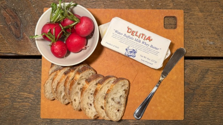 Delitia Buffalo Milk Butter in its packaging on a cutting board next to a butter knife, sliced baguette, and bowl of radishes
