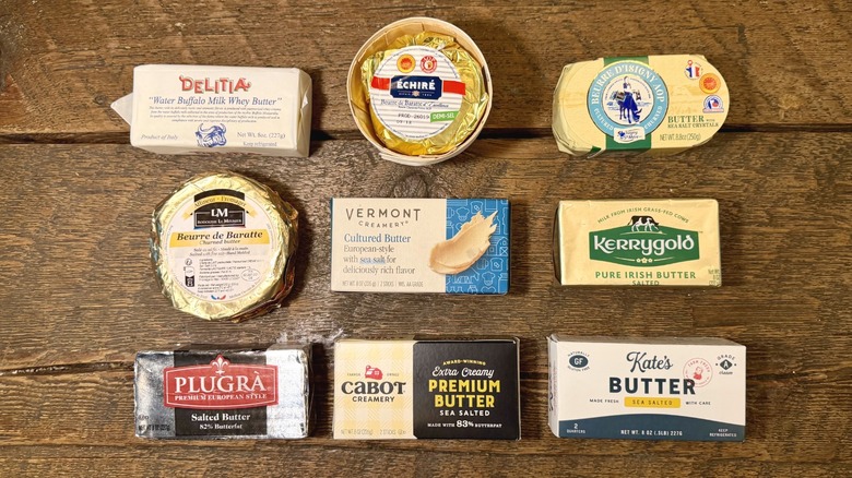 Nine premium butters on a wooden table, including Delitia, Echire, Isigny Ste Maire, Rodolphe le Meunier, Vermont Creamery Cultured Butter, Kerrygold, Plugra, Cabot Creamery Extra Creamy Premium Butter, and Kate's Butter