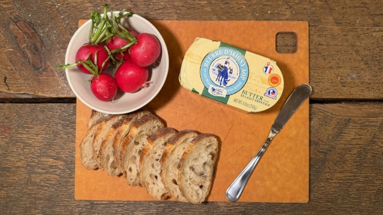 Isigny Ste Mère butter in its packaging on a cutting board next to a butter knife, sliced baguette, and bowl of radishes