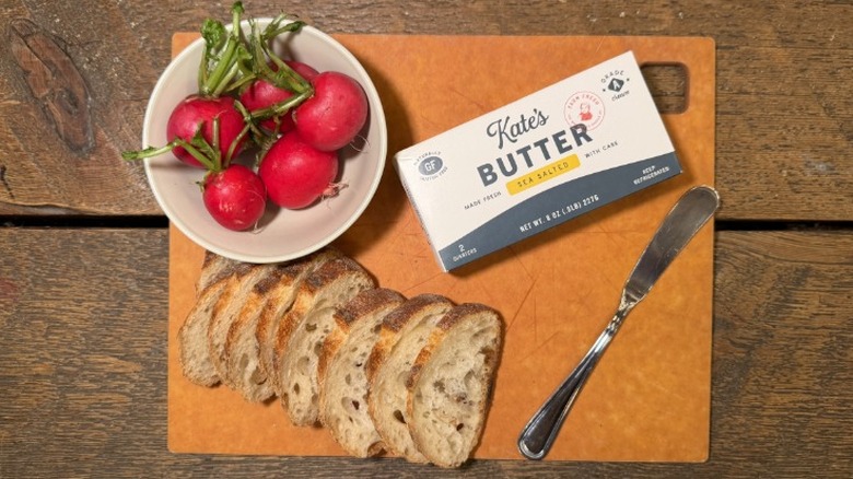 Kate's Butter in its packaging on a cutting board next to a butter knife, sliced baguette, and bowl of radishes