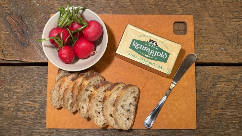 Kerrygold butter in its packaging on a cutting board next to a butter knife, sliced baguette, and bowl of radishes