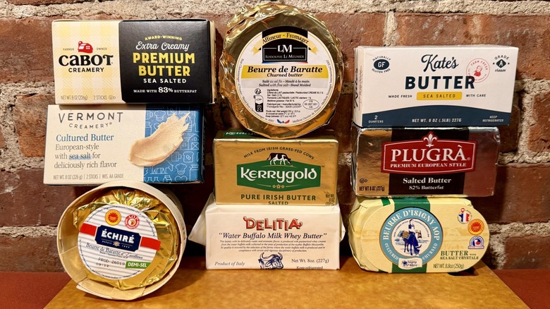 Nine premium butters against a brick wall, including Delitia, Echire, Isigny Ste Maire, Rodolphe le Meunier, Vermont Creamery Cultured Butter, Kerrygold, Plugra, Cabot Creamery Extra Creamy Premium Butter, and Kate's Butter