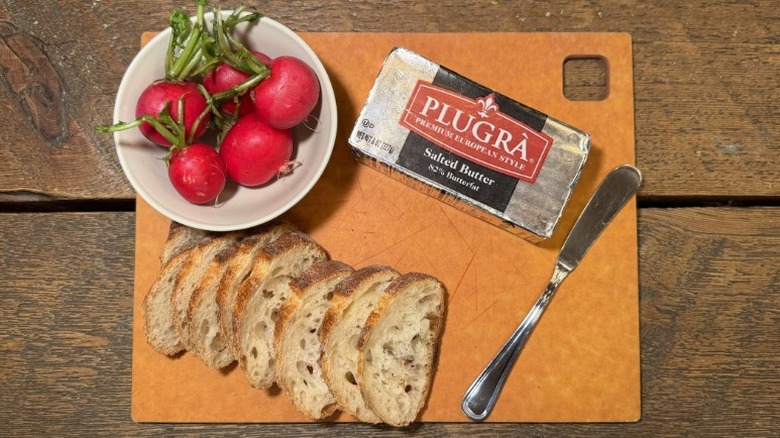 Plugra butter in its packaging on a cutting board next to a butter knife, sliced baguette, and bowl of radishes