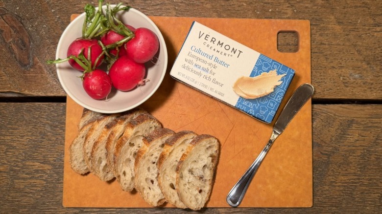 Vermont Creamery Cultured Butter in its packaging on a cutting board next to a butter knife, sliced baguette, and bowl of radishes