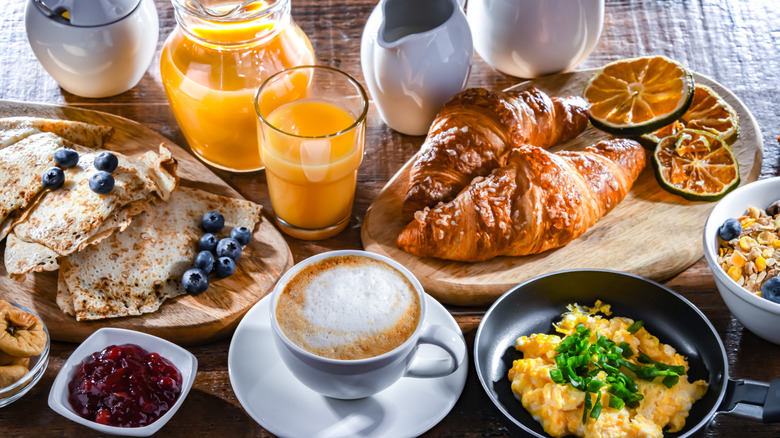 Variety of breakfast foods and beverages, such as croissants, eggs, latte, orange juice, jam, and more.