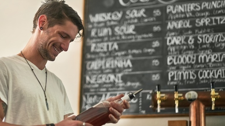 A smiling bartender holding a bottle with a drink menu behind him