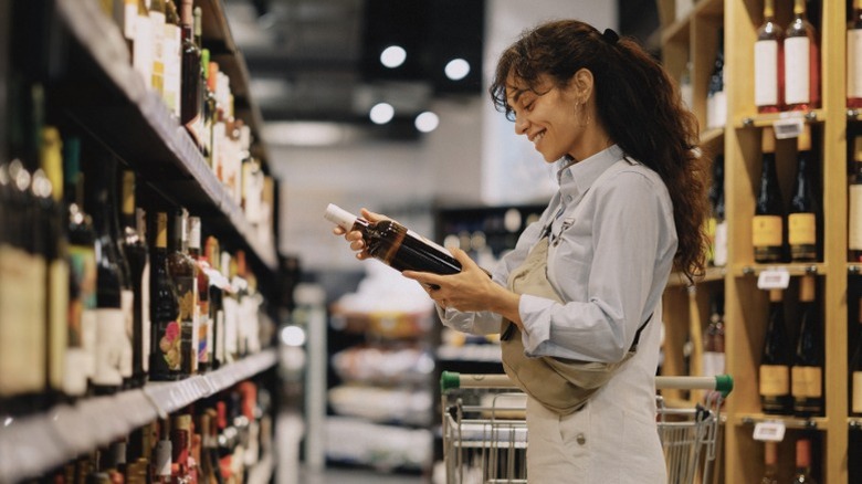A long-haired woman holding a wine bottle in a store aisle full of alcohol
