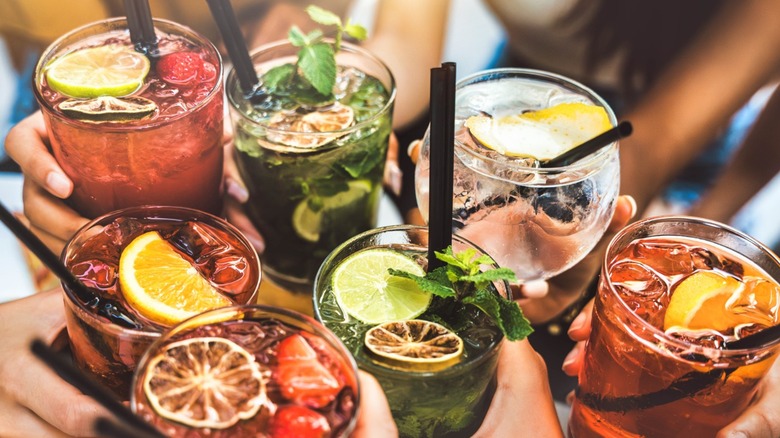 Several hands holding up seven cocktail glasses with garnish