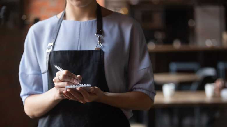 A waitress in a black apron holding a notepad and pen