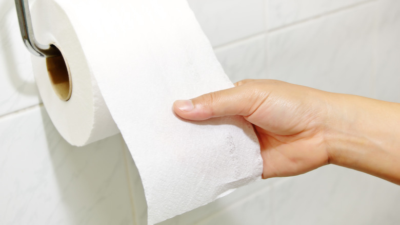 A hand pulling toilet paper off of a roll in front of a white backsplash