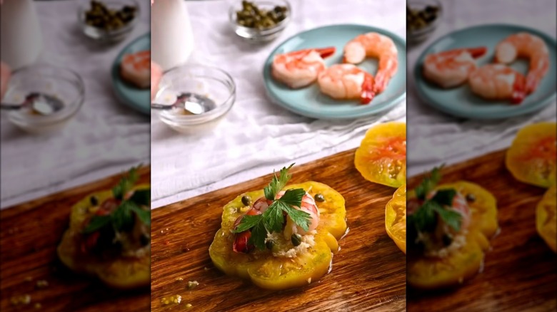 Shrimp cocktail-style tomato on a cutting board with shrimp, salt, and capers in the background