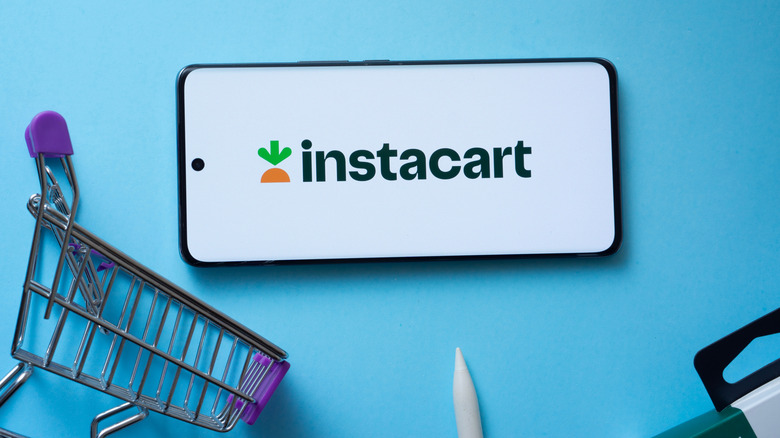 An Instacart logo on an iPhone next to a mini shopping cart and a pen